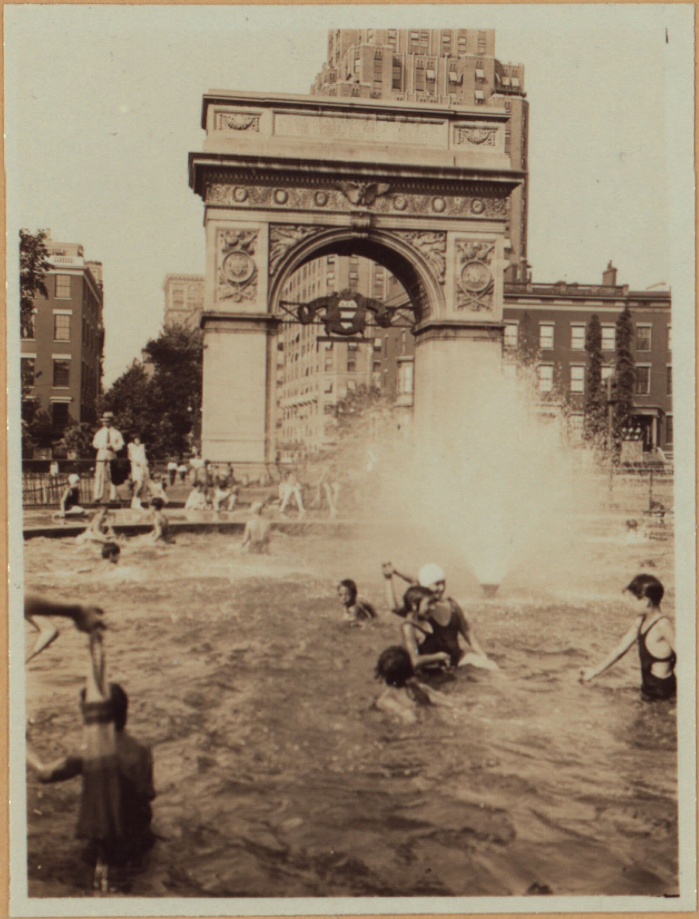 Manhattan: Washington Square North - 5th Avenue: Washington Square Park, showing the center fountain with the Washington Arch in the background, with children at play in the water. 18-22 Washington Square North, west of Fifth Avenue, are the houses in background.